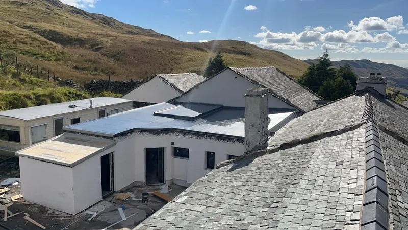 New grey flat roof on a white single-storey extension beside older slate roofs, with fells behind