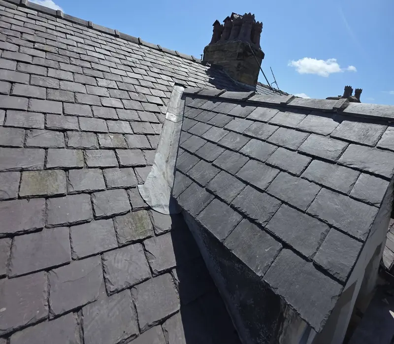 Natural slate roof with a lead-lined valley beside a small gable and a stone chimney stack against a blue sky