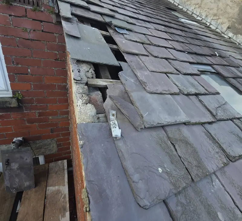 Natural slates slipped out of line at the edge of a roof beside a brick wall